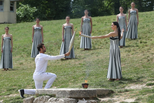 扮演最高女祭司的演員瑪麗·米娜（前右）點燃第一棒火炬手——希臘運動員、東京奧運會男子賽艇項目金牌得主斯特凡諾斯·恩圖斯科斯手中的火炬。新華社記者 趙丁喆 攝